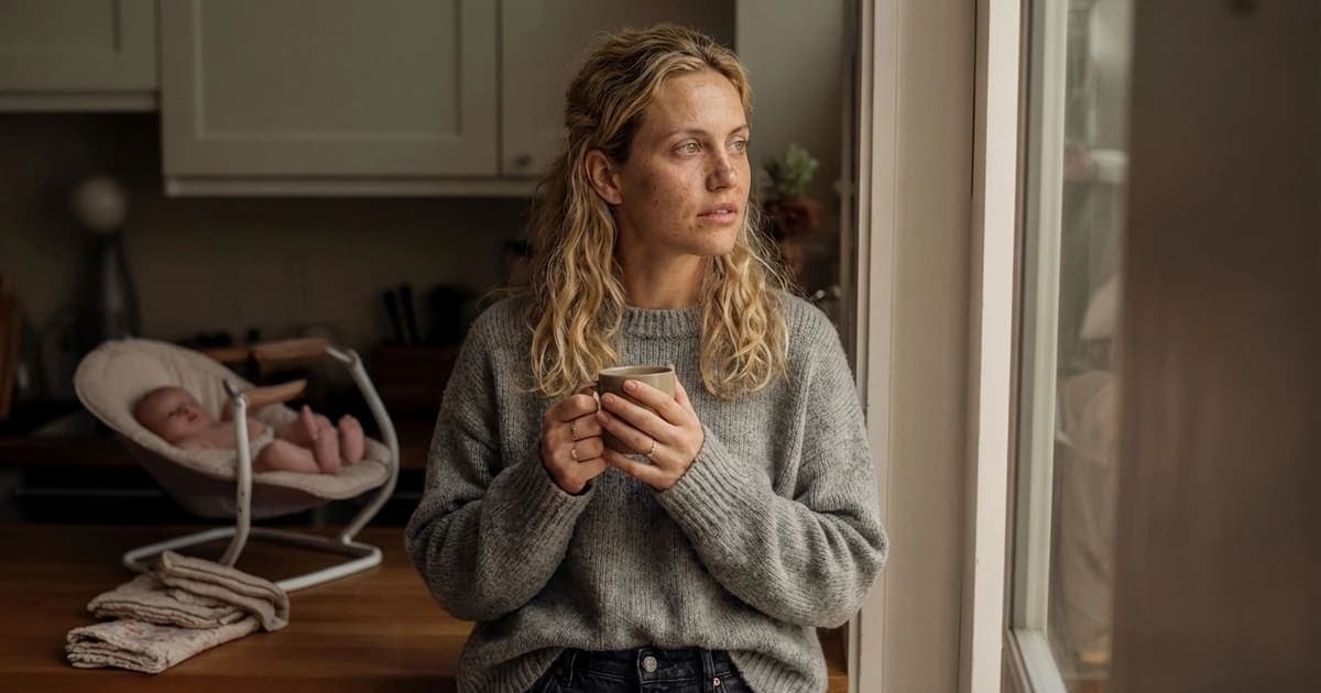 Postpartum mother standing in sunlit kitchen doorway holding warm cup of tea during fourth trimester recovery, natural golden hour light, editorial lifestyle photograph