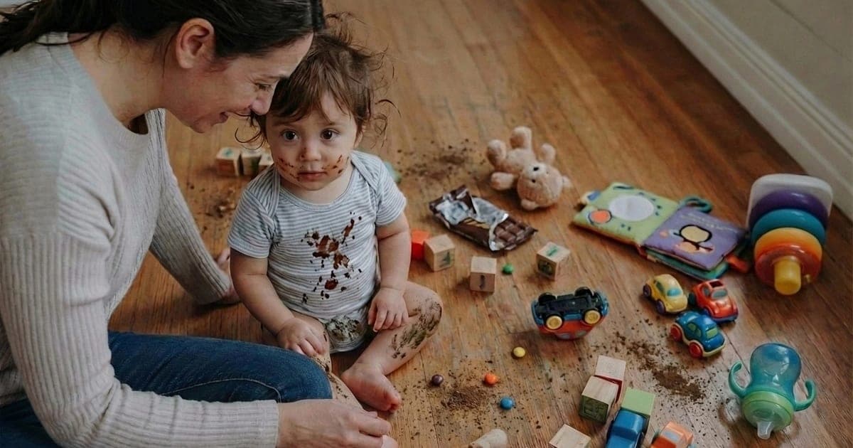 Mother sitting calmly on kitchen floor beside chocolate-covered toddler surrounded by scattered toys, demonstrating gentle parenting discipline without yelling