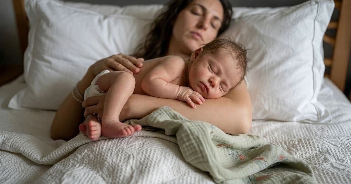 Newborn baby sleeping peacefully on mother's bare chest in soft morning light after a long cluster feeding session, with mother's hand gently cradling the baby's back.